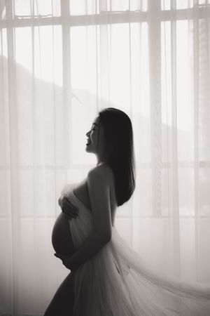 A silhouetted pregnant woman in a flowing dress stands by sheer curtains in a dreamy black and white maternity photo.一張夢幻般的黑白孕婦照中，一位身著飄逸長裙