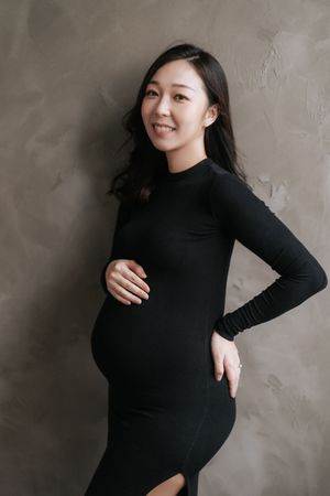 A series of maternity photos showing a woman in a black sweater posing against a gray textured wall.一組孕婦照，照片中一位身穿黑色毛衣的女子倚靠在灰色紋理牆前擺姿勢。