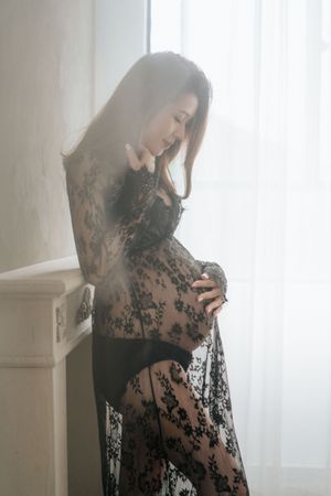 A pregnant woman in a sheer black lace dress poses by a window in soft natural lighting一位身穿黑色薄紗蕾絲連身裙的孕婦在柔和的自然光線下，倚窗擺姿勢拍照。