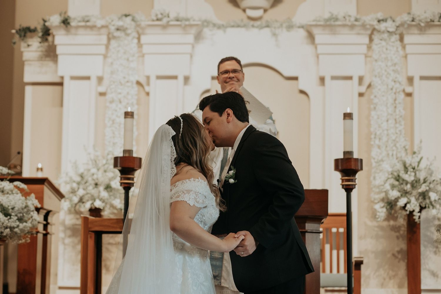 Bride and groom share their first kiss at the altar during a church wedding ceremony with floral decorations.