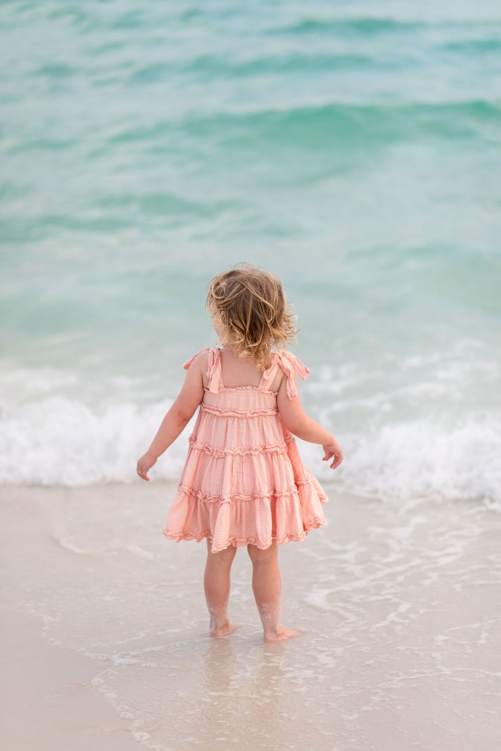Toddler girl in pink ruffle dress standing at ocean shoreline with turquoise waves at the beach.