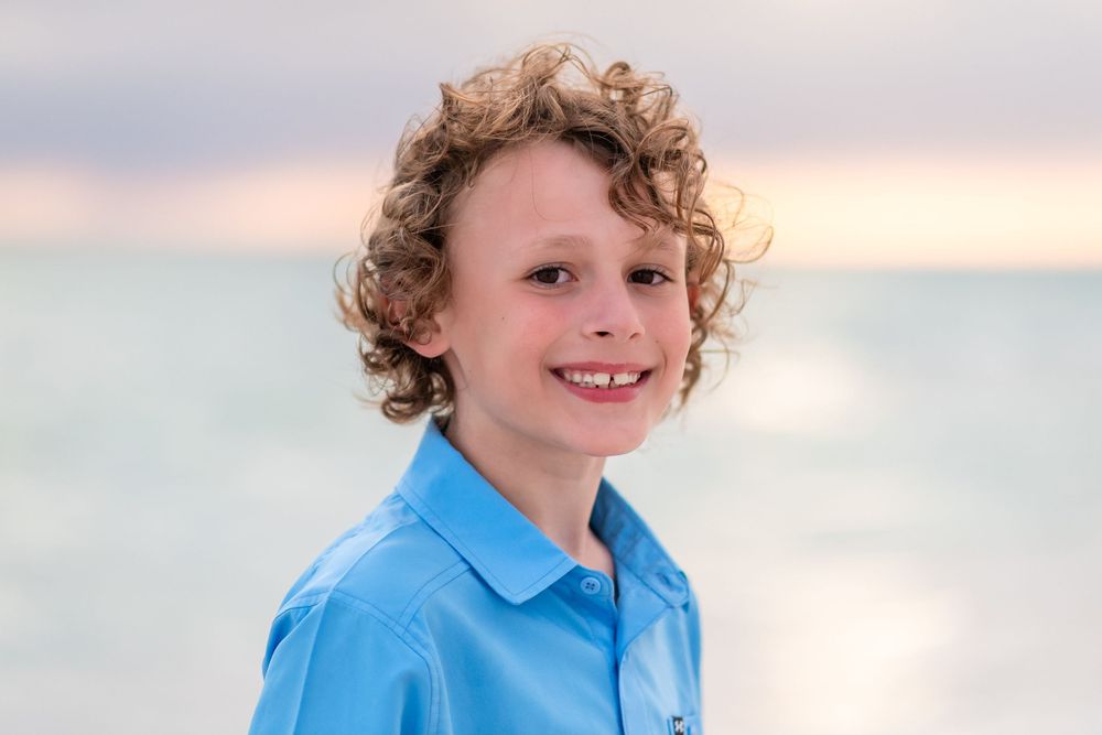 Smiling boy with curly hair wearing a blue polo shirt at the beach during sunset.