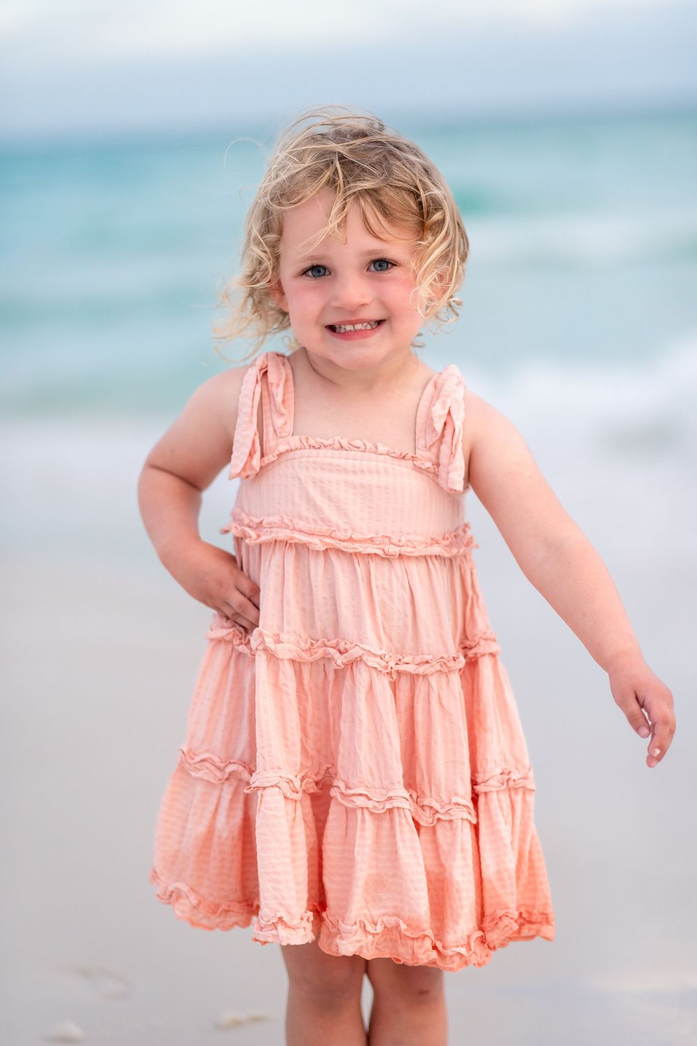 Smiling toddler girl with curly blonde hair wearing a pink ruffle dress standing on a sandy beach.