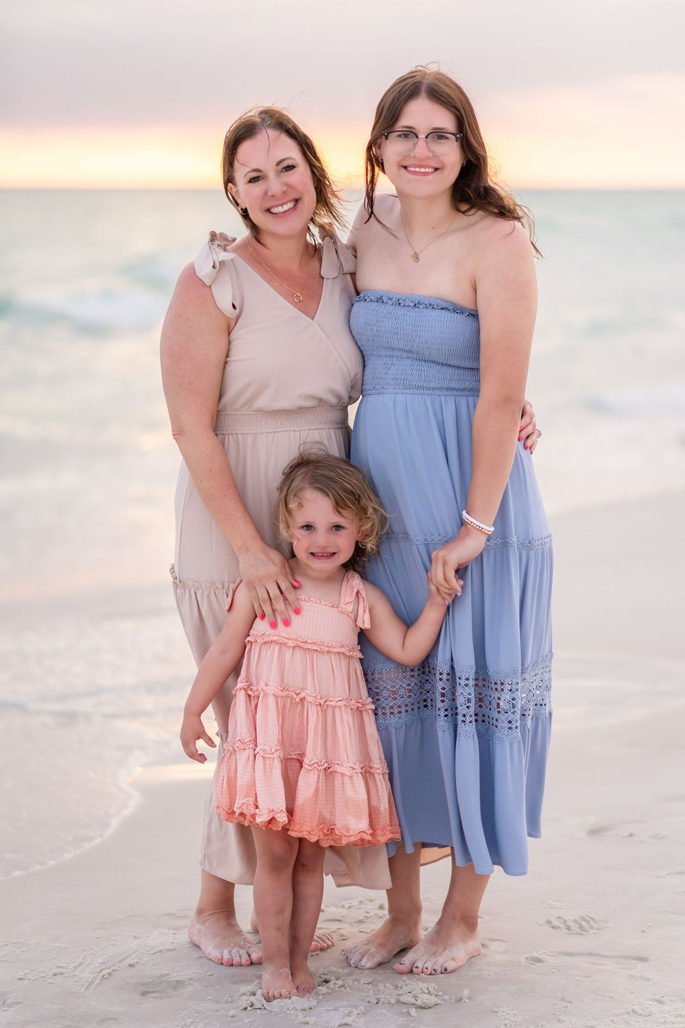 Three generations of women pose together on a sandy beach at sunset, wearing pastel dresses.