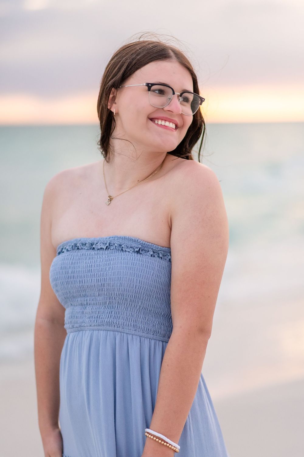 Smiling woman in blue strapless dress and glasses posing on beach at sunset with ocean behind her.