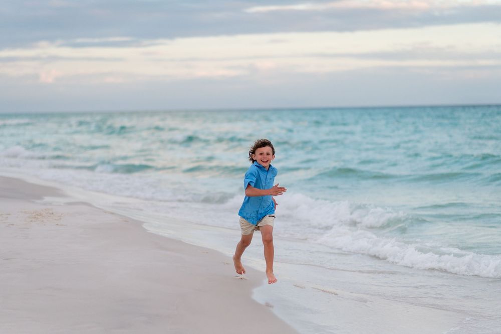 A happy child runs barefoot along a sandy beach with turquoise ocean waves under a cloudy sky.