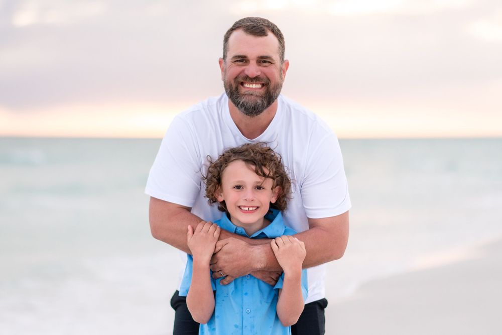A smiling father hugs his curly-haired son on a sandy beach at sunset, both wearing casual attire.