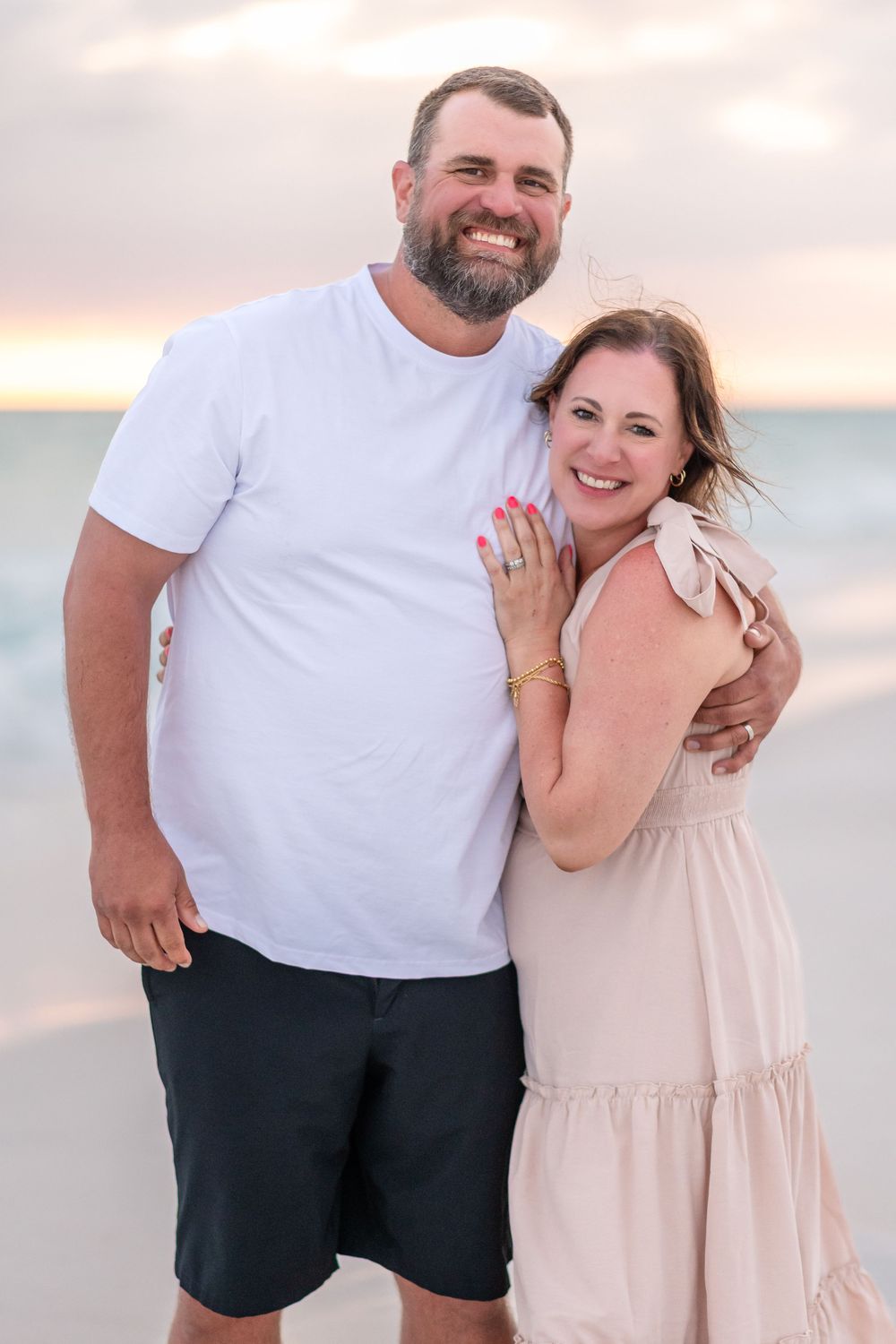 Smiling couple embracing on a beach at sunset, man in white t-shirt and dark shorts, woman in light pink dress.