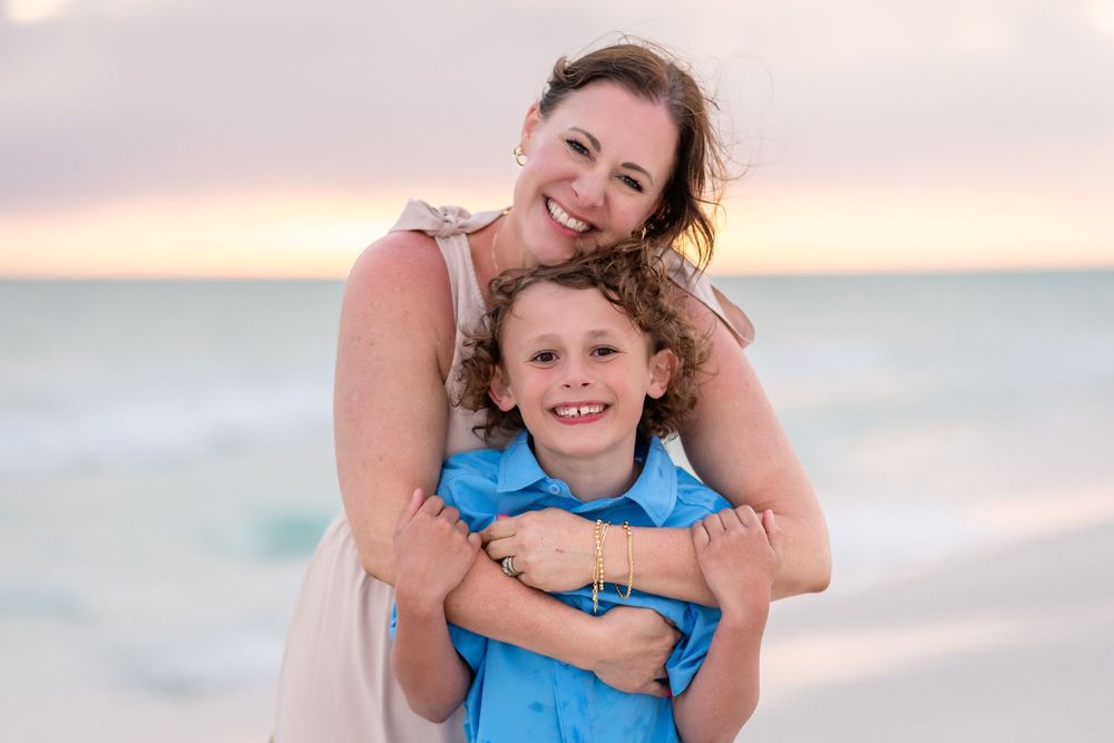 A smiling woman hugs a young curly-haired boy in a blue shirt on a beach at sunset.