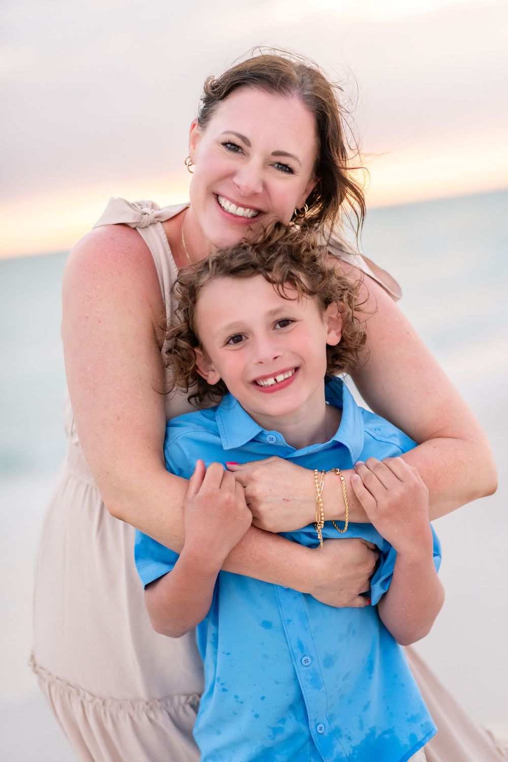 A smiling mother hugs her curly-haired boy on a beach at sunset, both wearing blue.