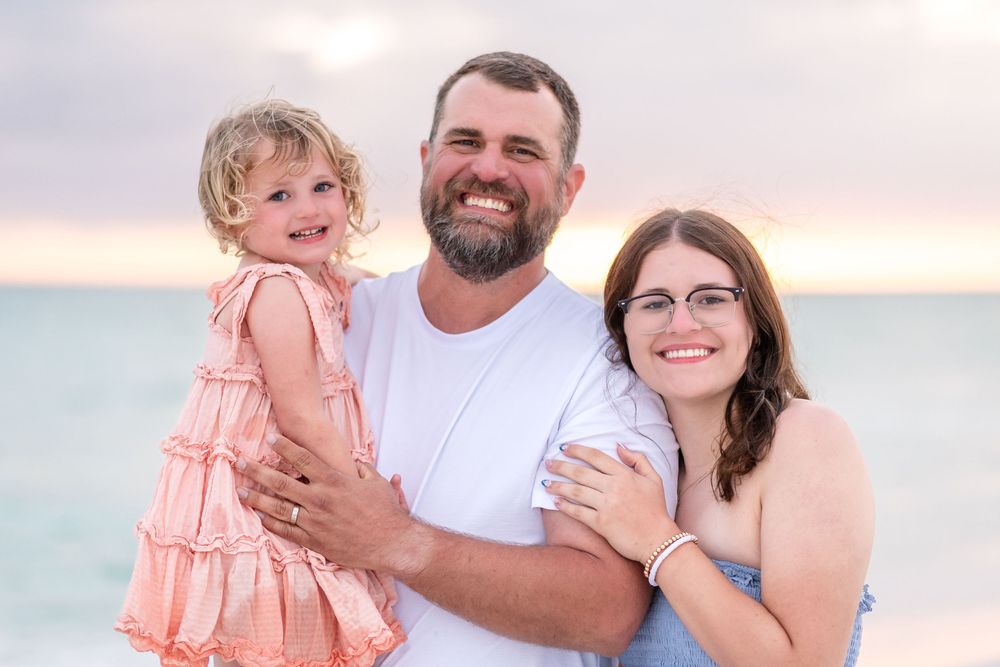 A smiling family of three poses on a beach at sunset, with a man holding a young girl in a pink dress beside a woman.