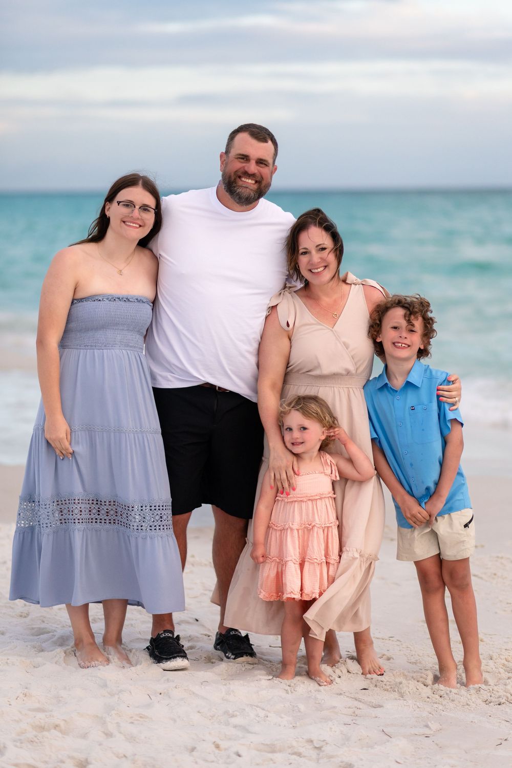 Family of five smiling together on a white sand beach with turquoise ocean waves in the background.