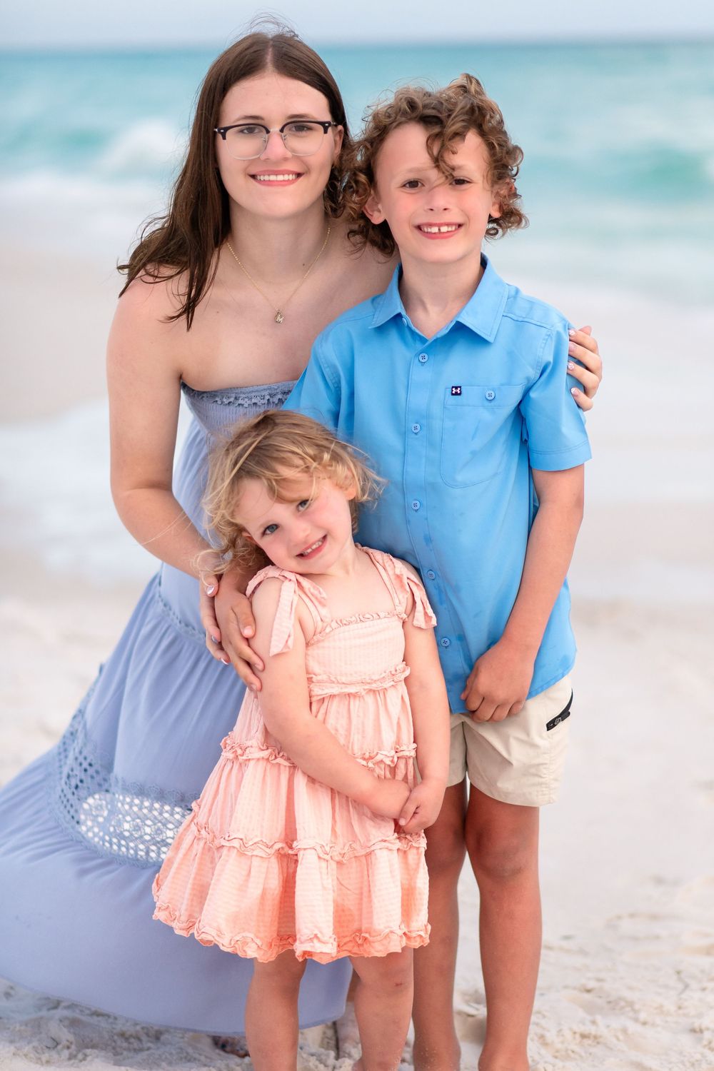 Woman in blue dress with two children smiling on a sandy beach with turquoise ocean waves in background.