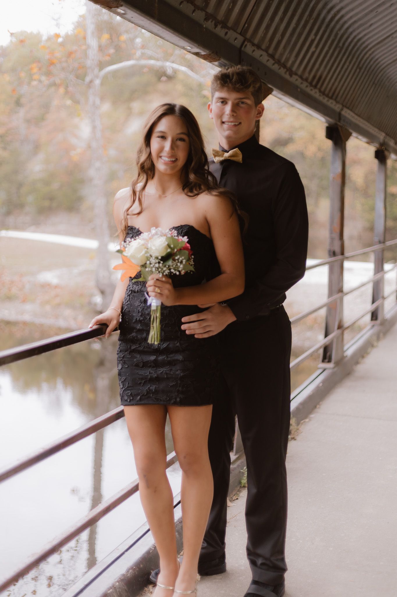 Couple in black formal attire posing on a covered bridge holding a white floral bouquet.