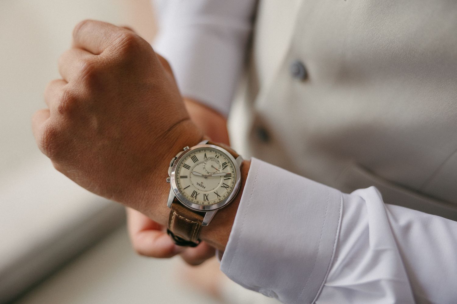 Man in white dress shirt and gray suit adjusting a classic silver Roman numeral wristwatch on his wrist.