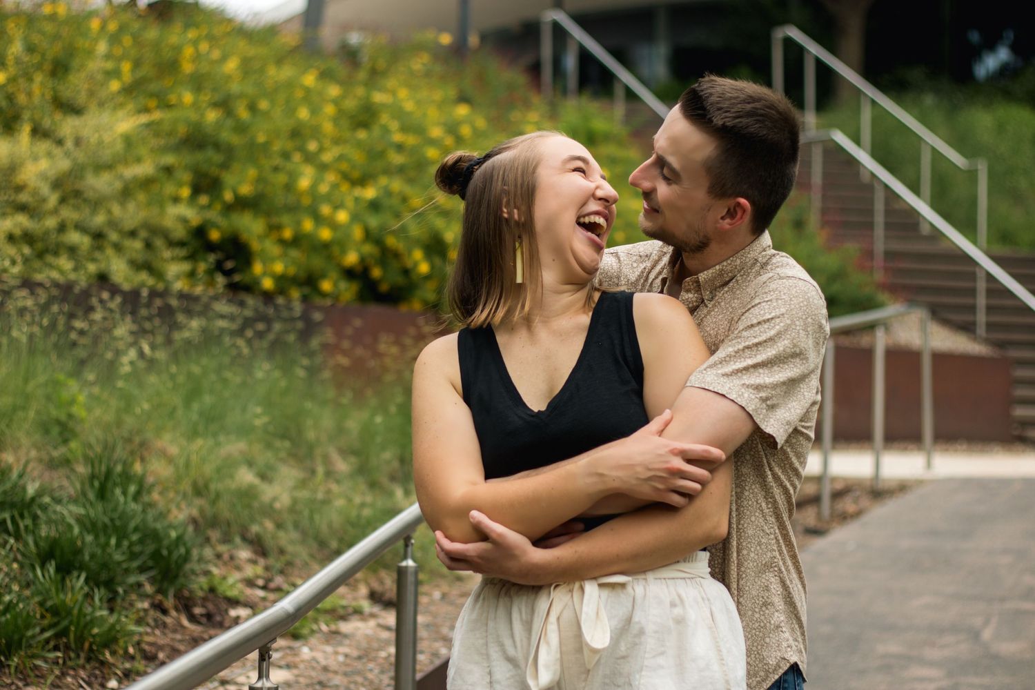 A laughing couple embraces outdoors near stairs and lush greenery on a sunny day.