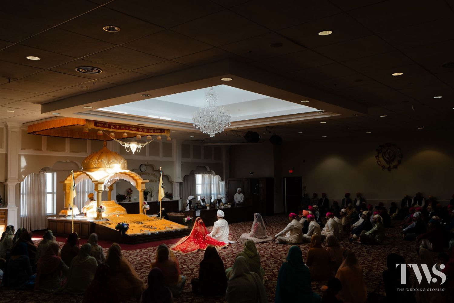 Elegant Sikh wedding ceremony in a grand ballroom with golden mandap and guests seated around the sacred space.