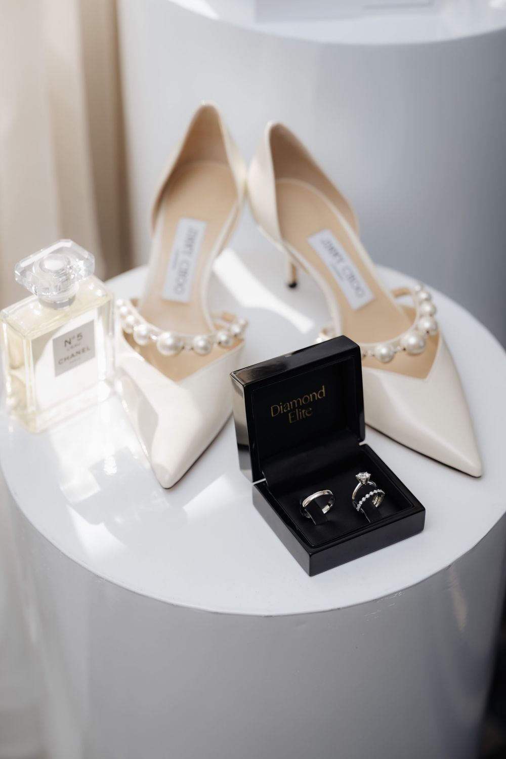 Bridal flat lay featuring Jimmy Choo pearl heels, Chanel No.5 perfume, and diamond wedding rings in a black box.