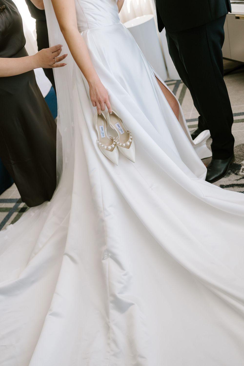 Bride in white satin gown holding pearl-embellished wedding shoes with flowing train and wedding party in background.
