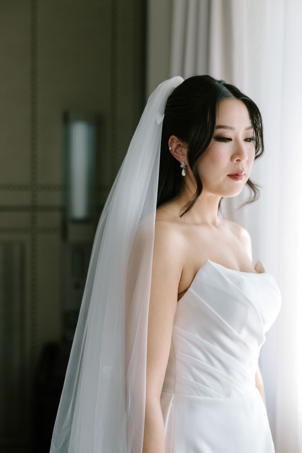 Elegant bride in strapless white gown and flowing veil gazes down by a window in soft natural light.