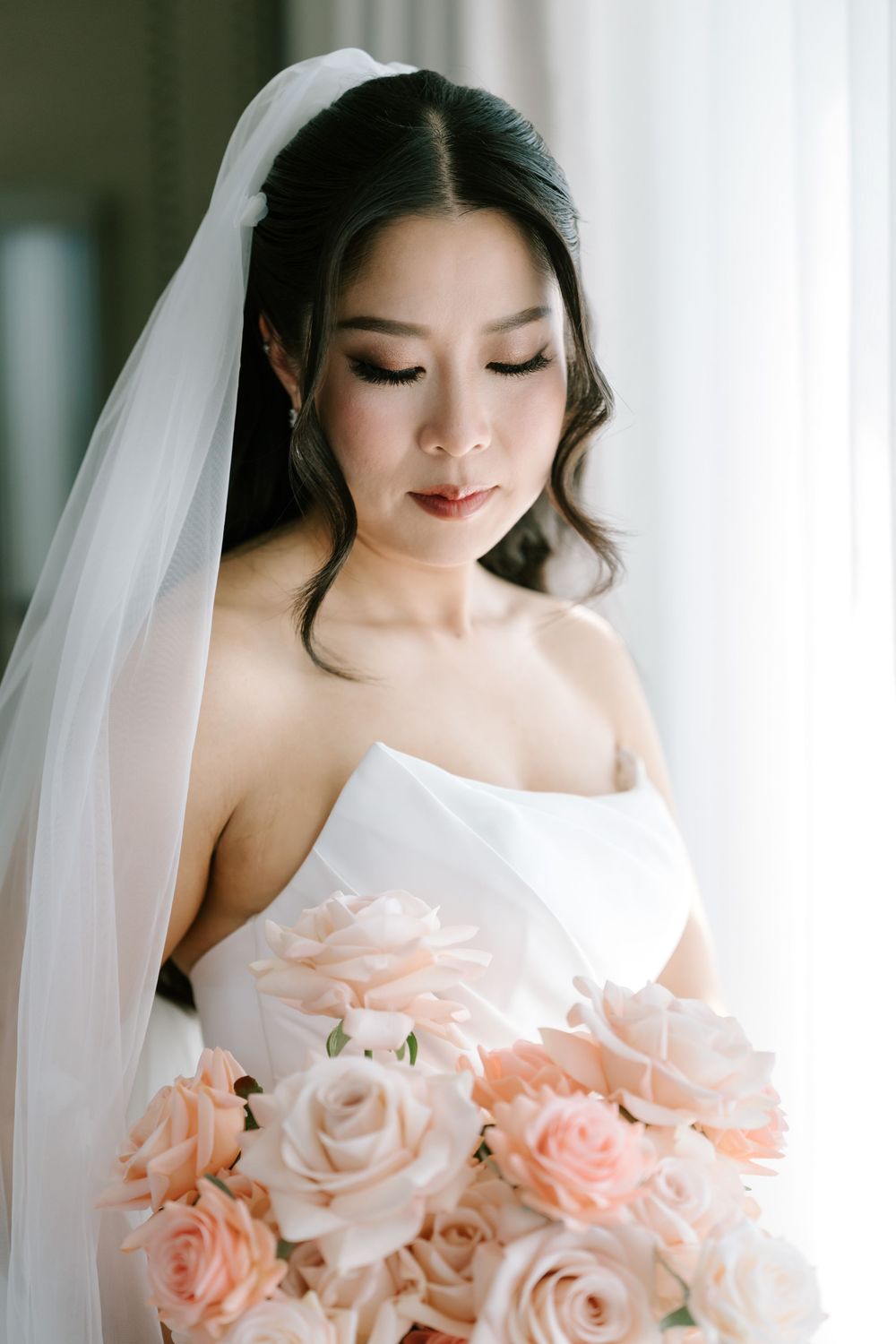 Asian bride in strapless white gown and veil gazes down at blush pink rose bouquet in elegant bridal portrait.