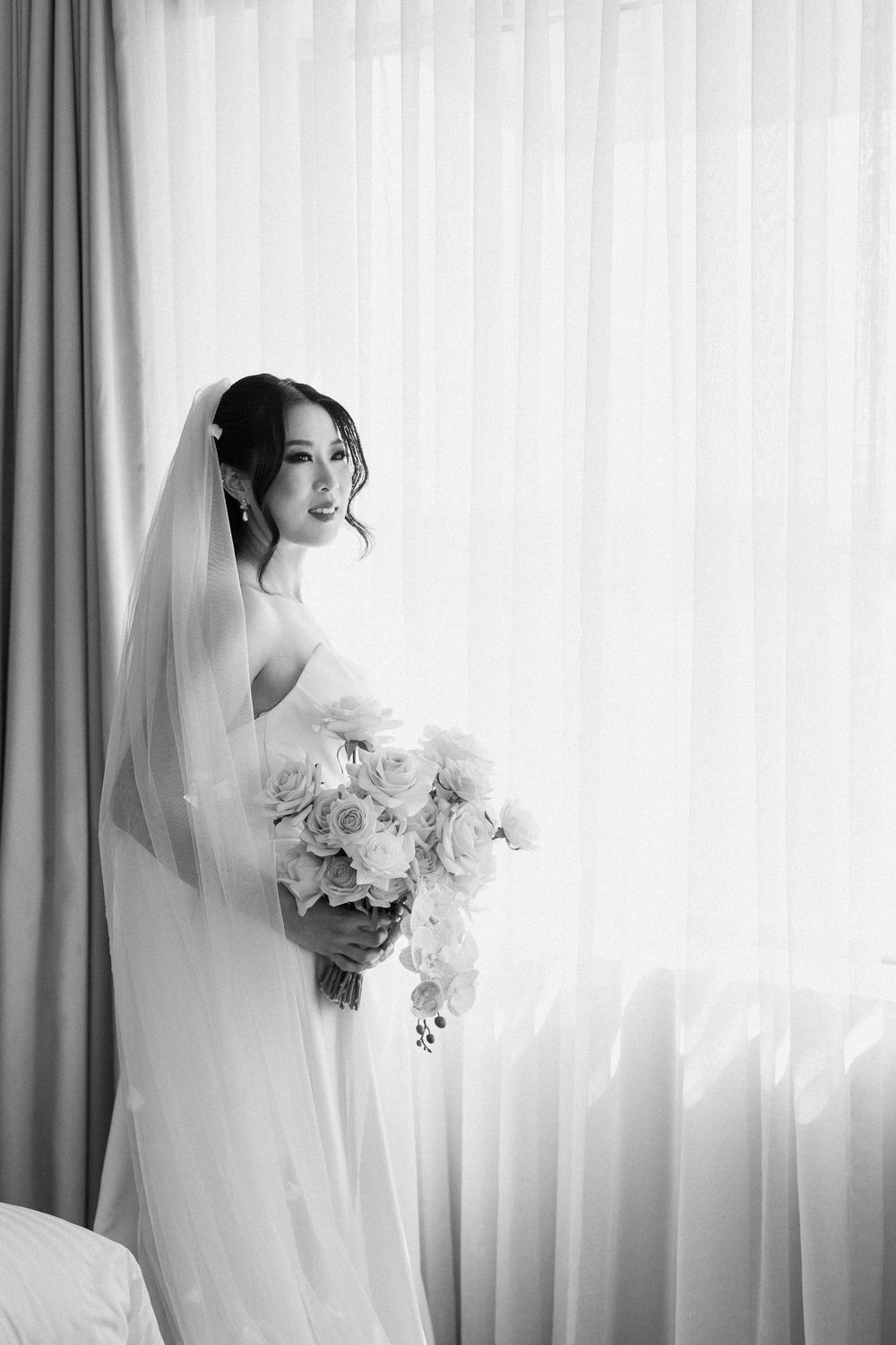Black and white photo of a bride in a white gown and veil holding a bouquet of roses near a bright window.