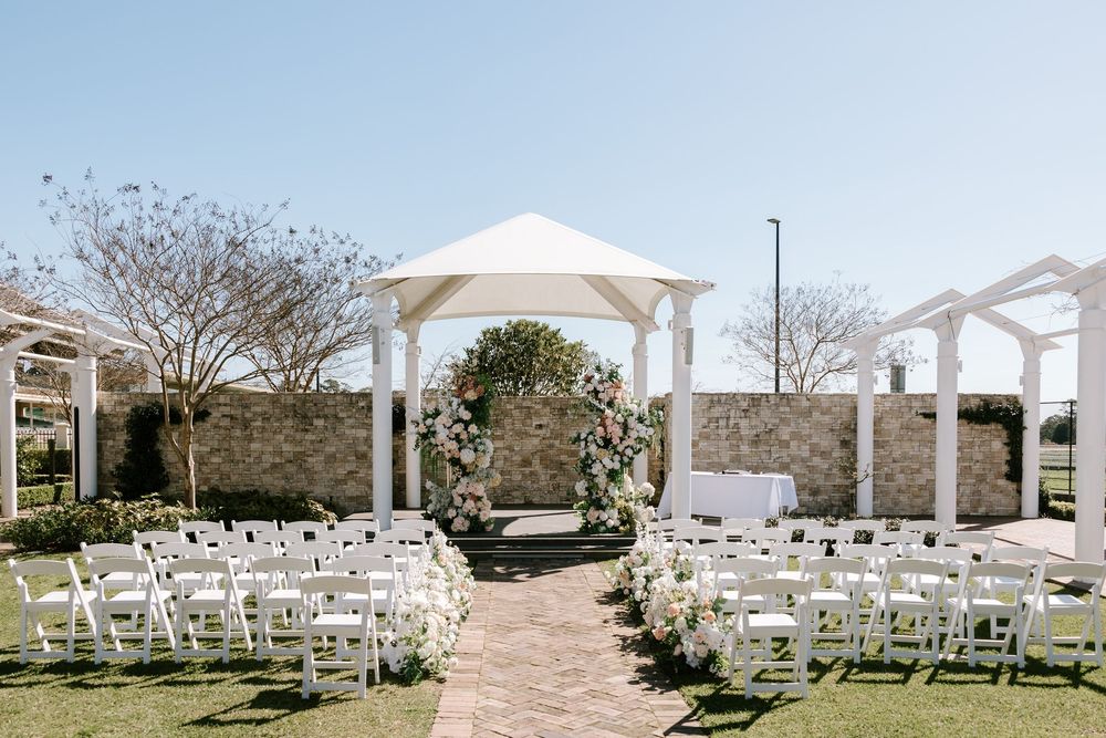 Elegant outdoor wedding ceremony setup with white chairs, floral arch, and white gazebo on a sunny day.