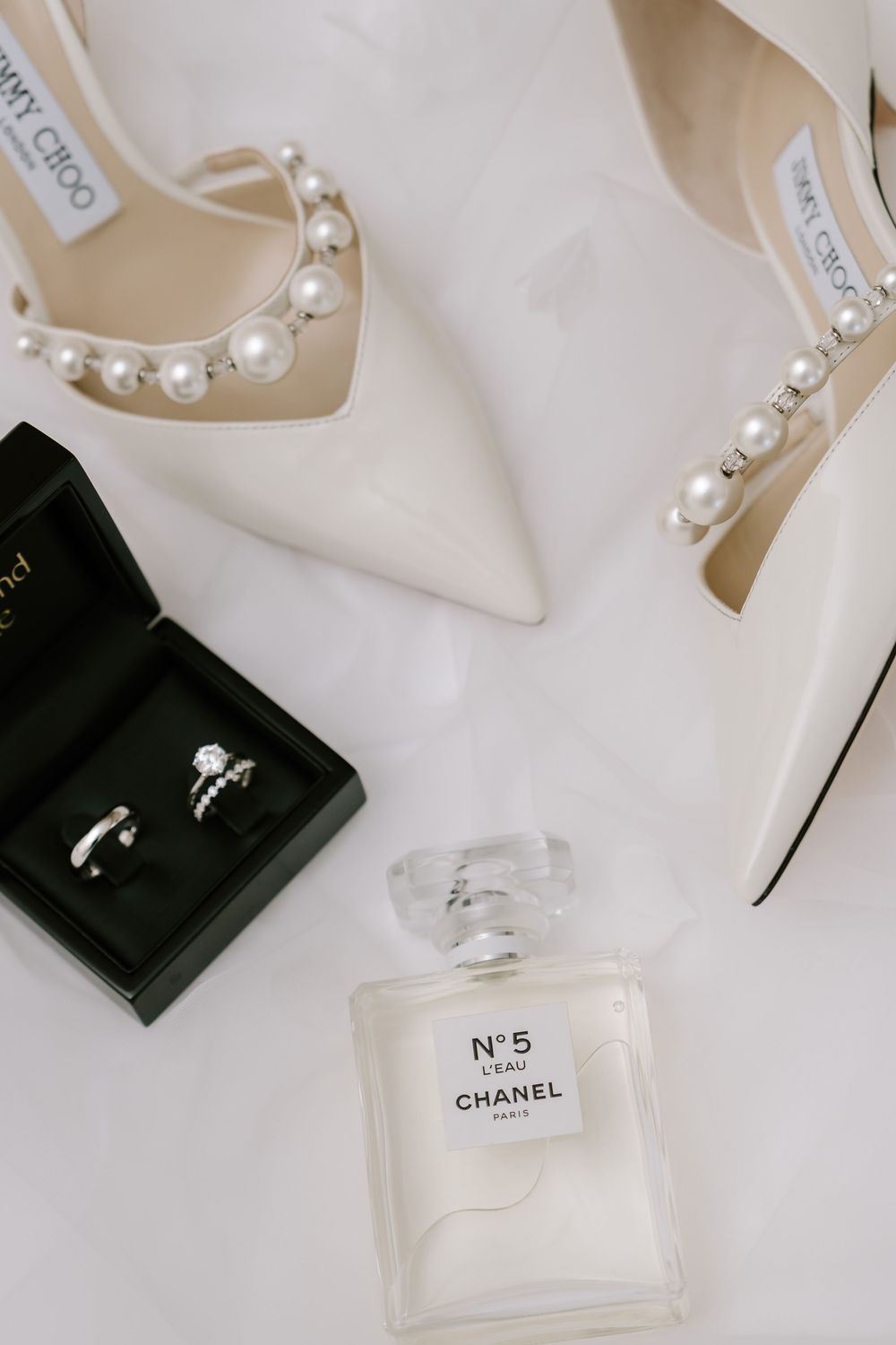 White Jimmy Choo pearl heels, Chanel No.5 L'Eau perfume, and diamond rings in a box on white fabric.