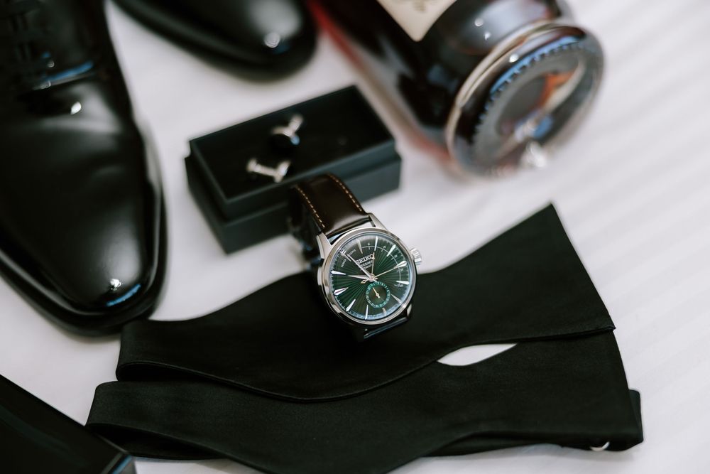 Elegant men's accessories flat lay featuring a green dial watch, black bow tie, leather shoes, and cologne bottle.