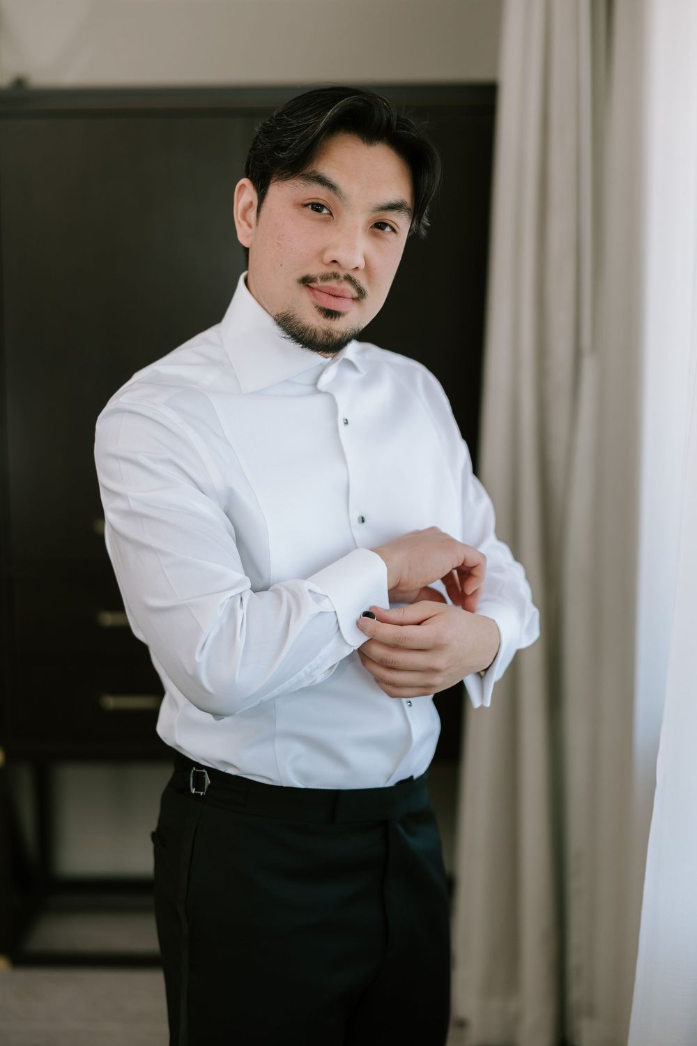 A groom in a white dress shirt adjusts his cufflinks while getting ready for his wedding day.