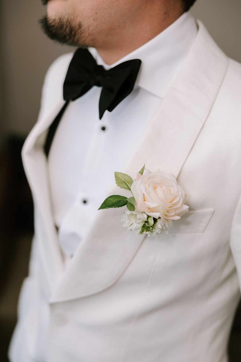 A groom wearing a white tuxedo jacket with black bow tie and blush rose boutonniere at a wedding.