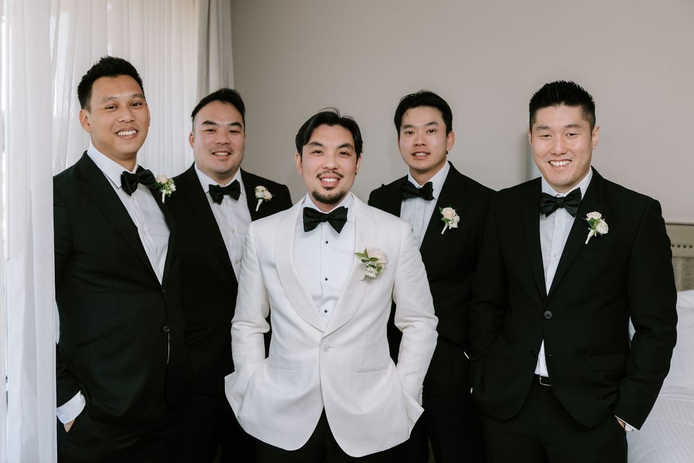 Groom in white tuxedo jacket poses with four groomsmen in black tuxedos and bow ties at wedding.