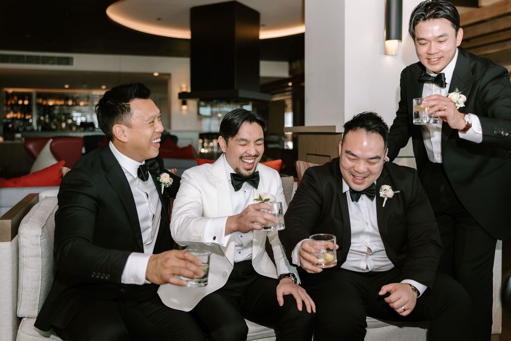 Four groomsmen in tuxedos laughing and toasting with drinks at a wedding reception venue.