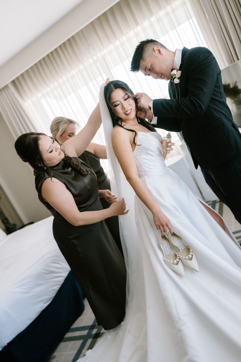 Bride in white gown gets veil adjusted by groom and bridesmaids in hotel room before wedding ceremony.
