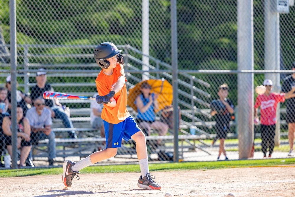A young baseball player in an orange jersey and blue shorts swings a bat during a sunny youth baseball game.