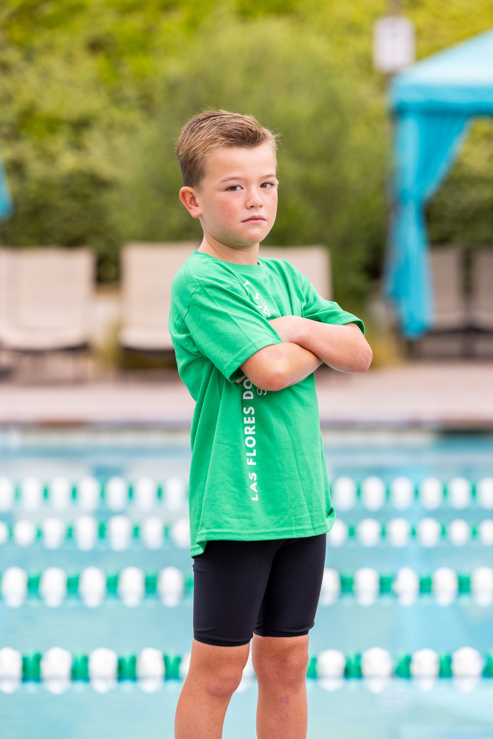 Young boy in green Las Flores Dolphins swim team shirt stands confidently with arms crossed beside a pool.