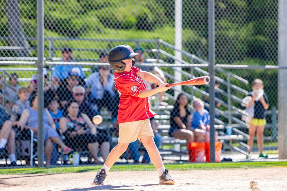 A young baseball player in a red uniform swings a bat during a sunny youth baseball game with spectators watching.