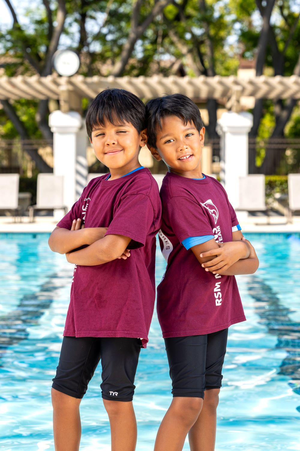 Two young boys in matching maroon swim team shirts stand back-to-back with arms crossed in front of a pool.