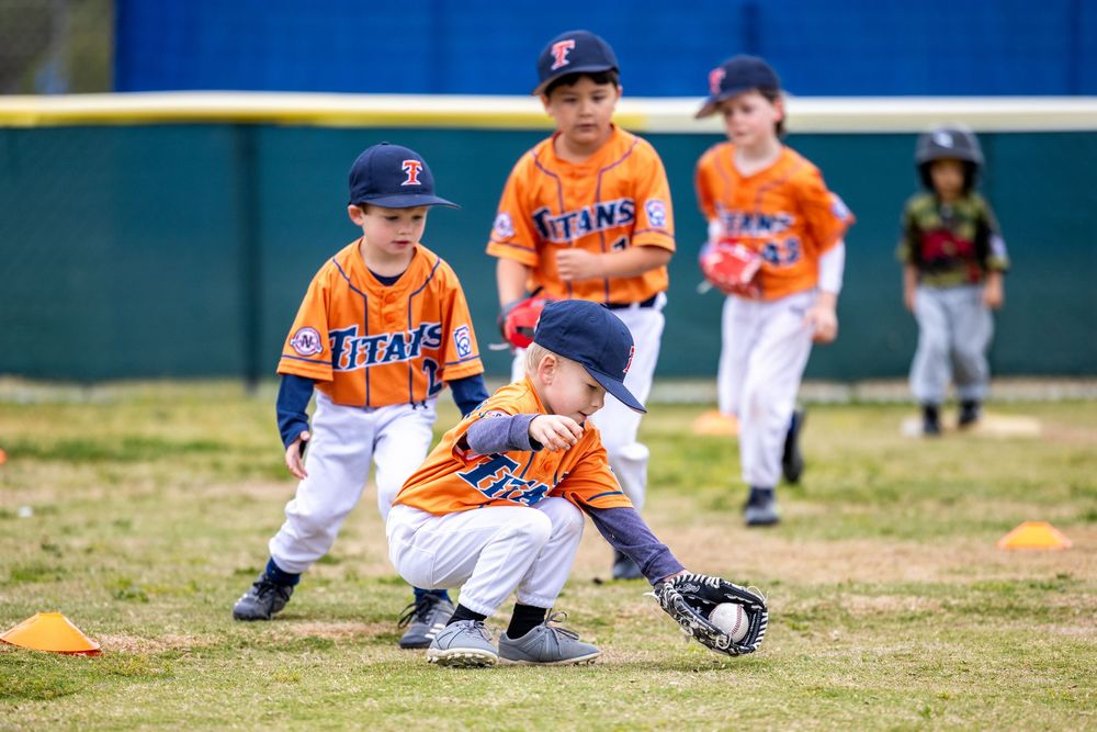 Young children in orange Titans baseball uniforms practice fielding during a youth baseball game on a green field.