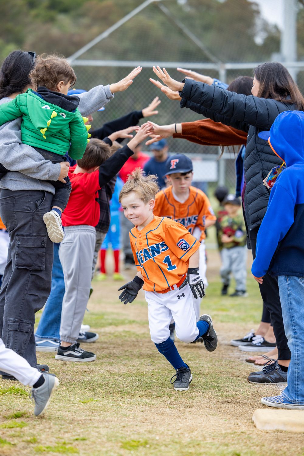 Smiling child in orange Titans baseball uniform runs through a high-five tunnel of cheering parents at a youth game.