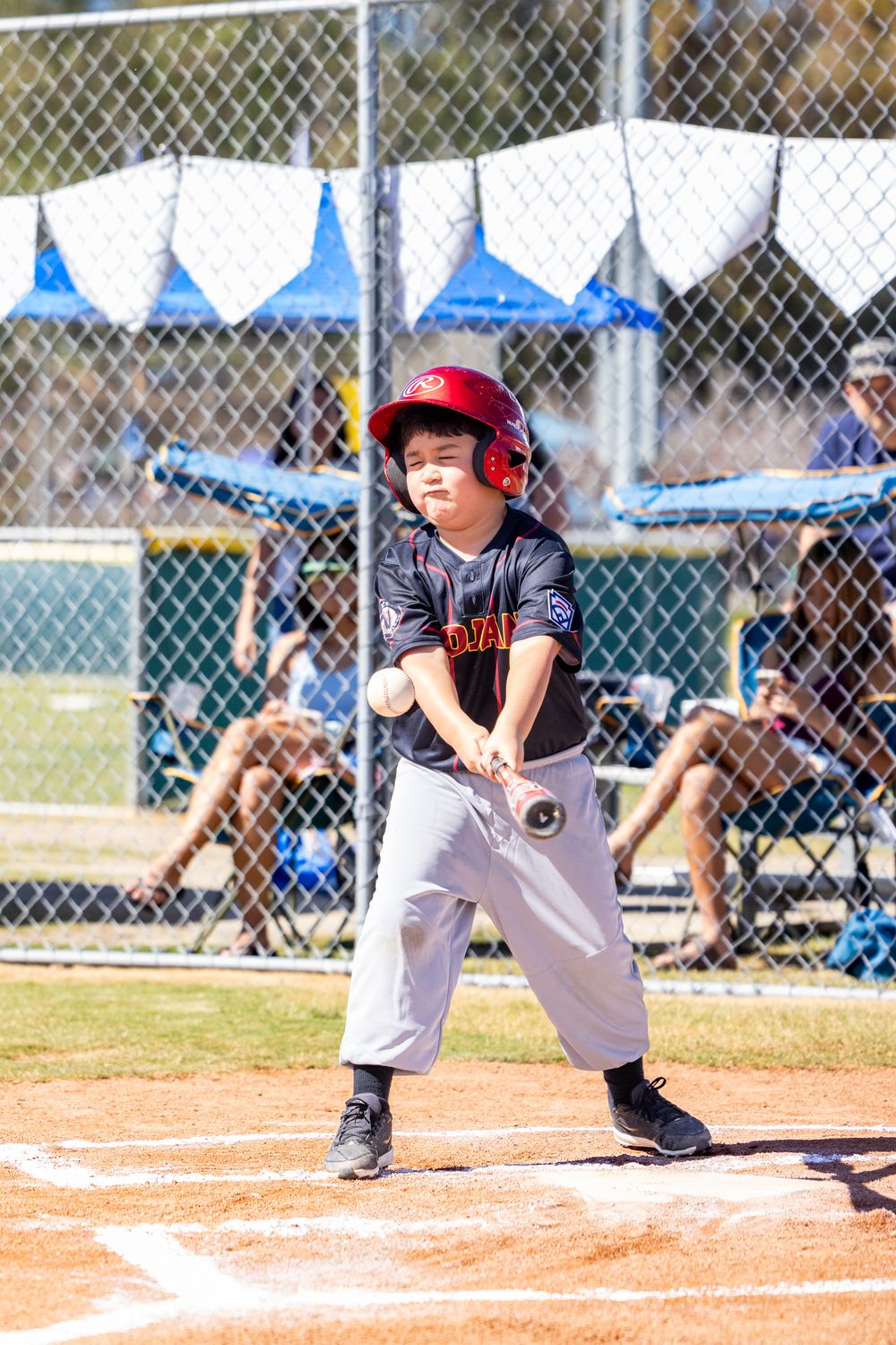 A young baseball player in a dark uniform and red helmet swings at a pitch during a youth baseball game.