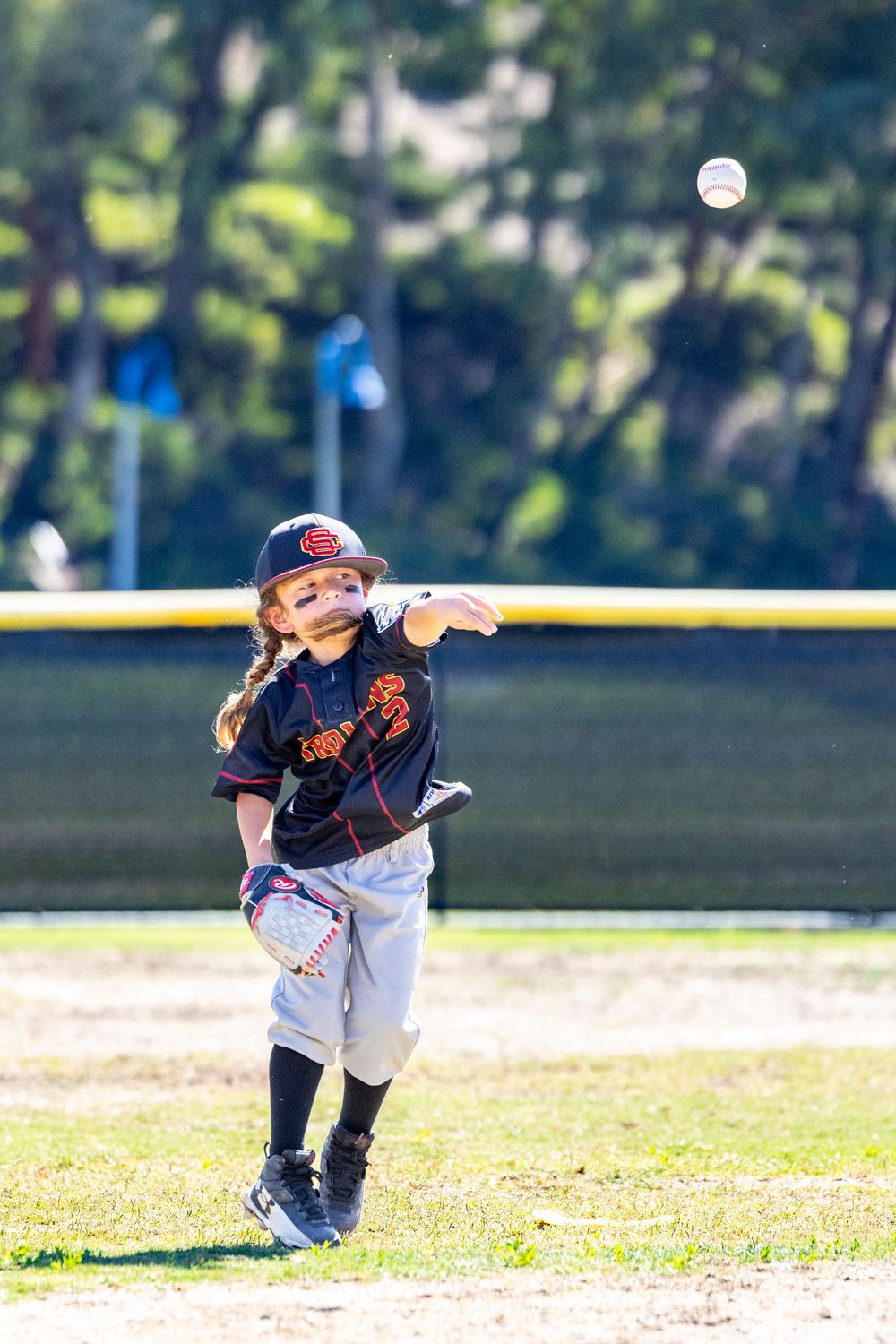 Young girl in black Trojans baseball uniform winds up to throw a ball during a youth baseball game outdoors.