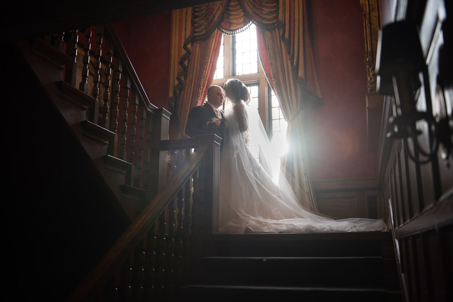 Dramatic silhouette of bride and groom on staircase with backlit window in historic venue.