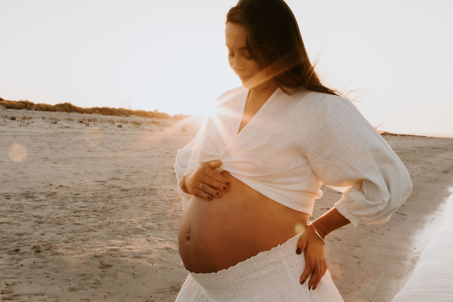 Pregnant woman in white outfit cradling her baby bump on a sunny beach at golden hour.