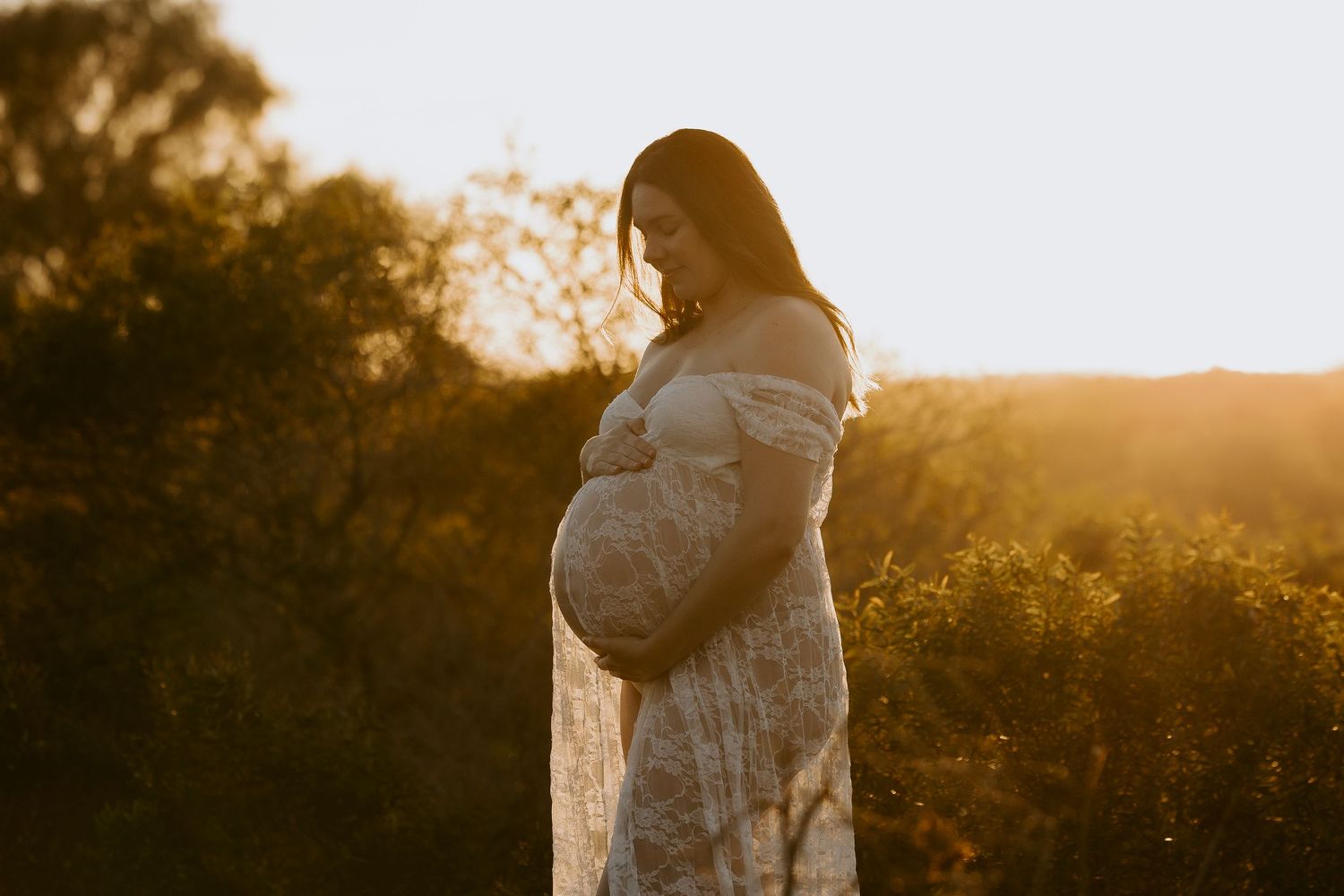Pregnant woman in white outfit cradling her baby bump on a sunny beach at golden hour.