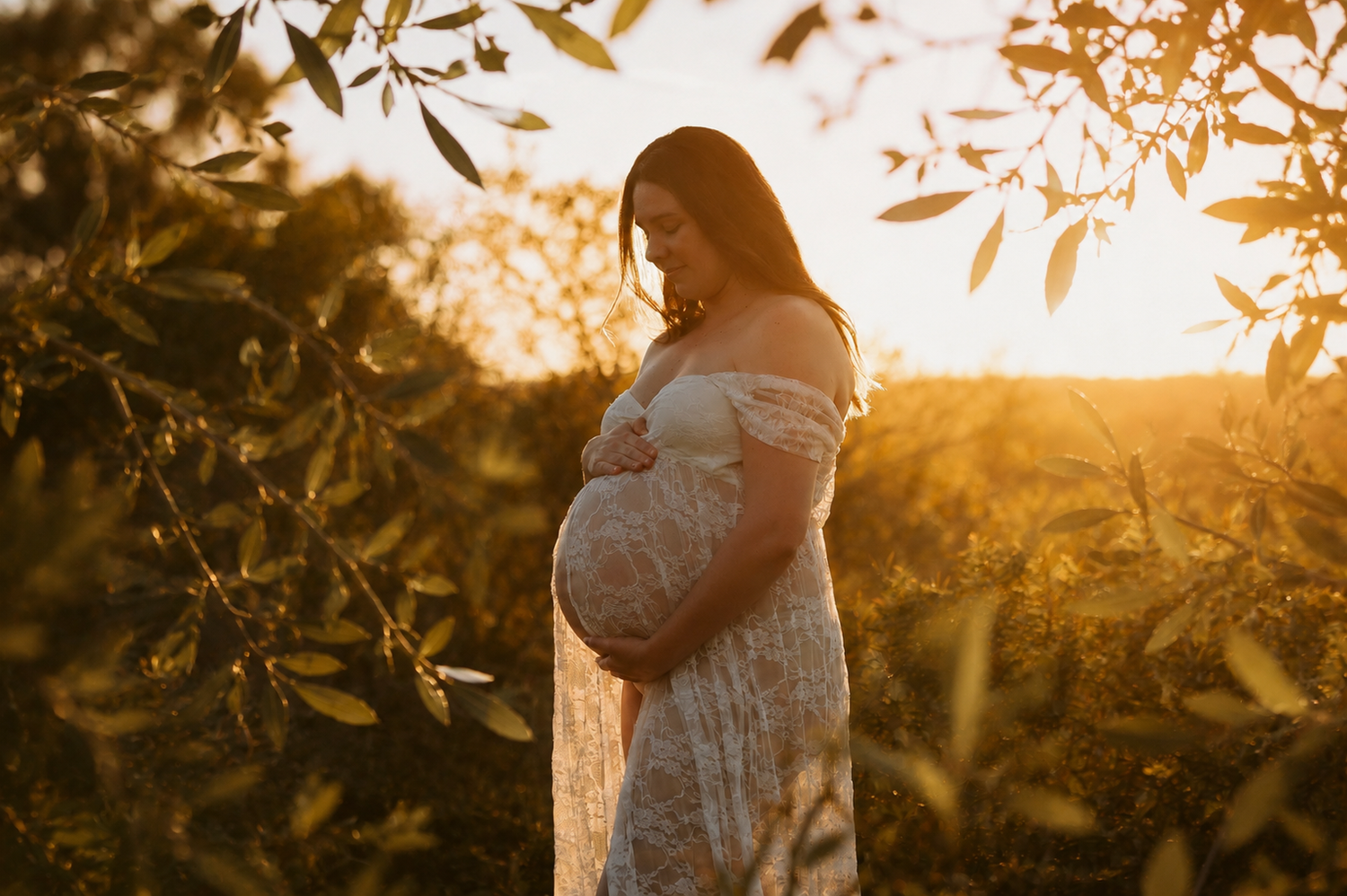 Pregnant woman in white outfit cradling her baby bump on a sunny beach at golden hour.