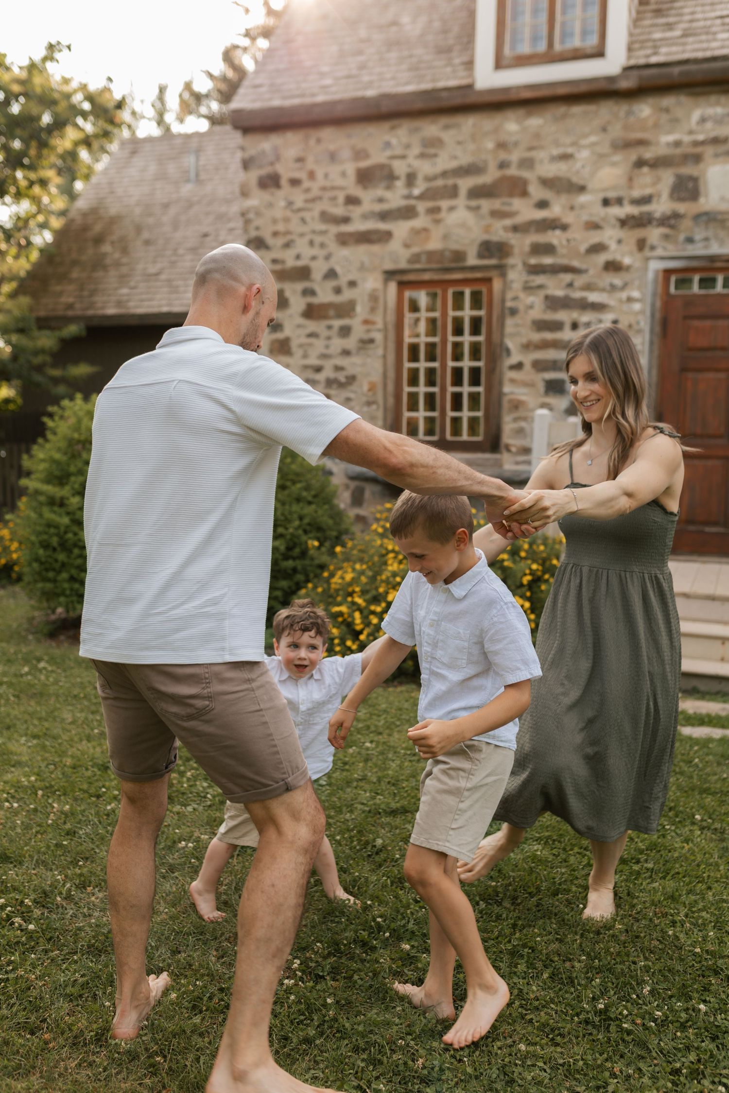 Joyful family dancing barefoot on green lawn outside rustic stone house in warm summer sunset.