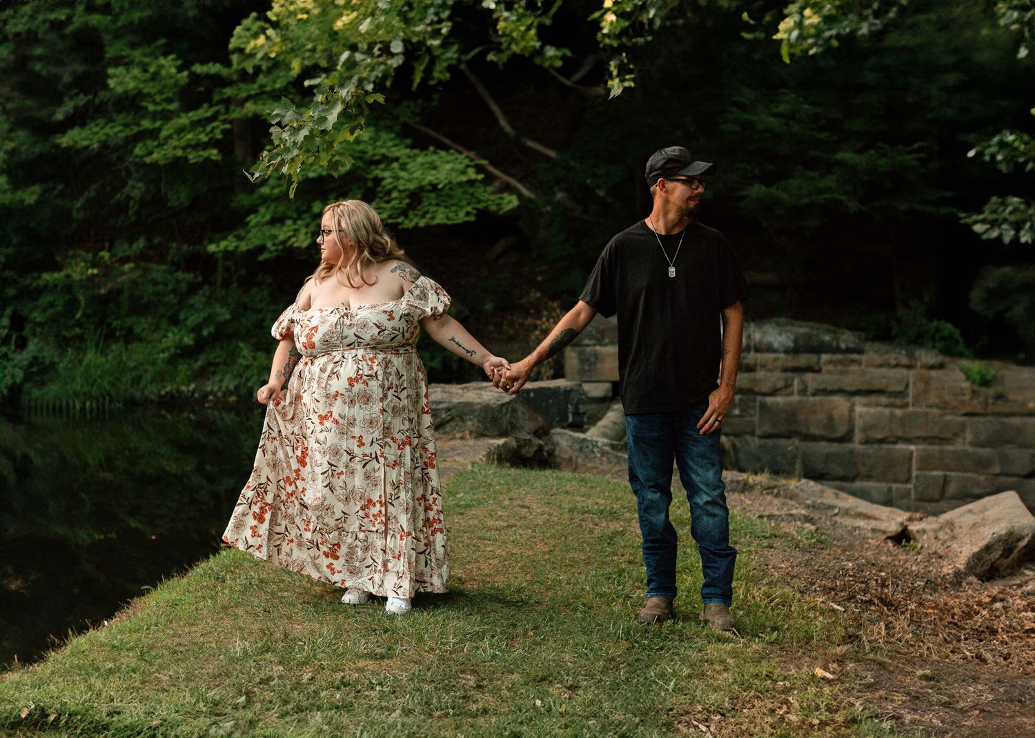 Pregnant woman in floral dress holds hands with man in black shirt and jeans near a creek in a lush green forest.