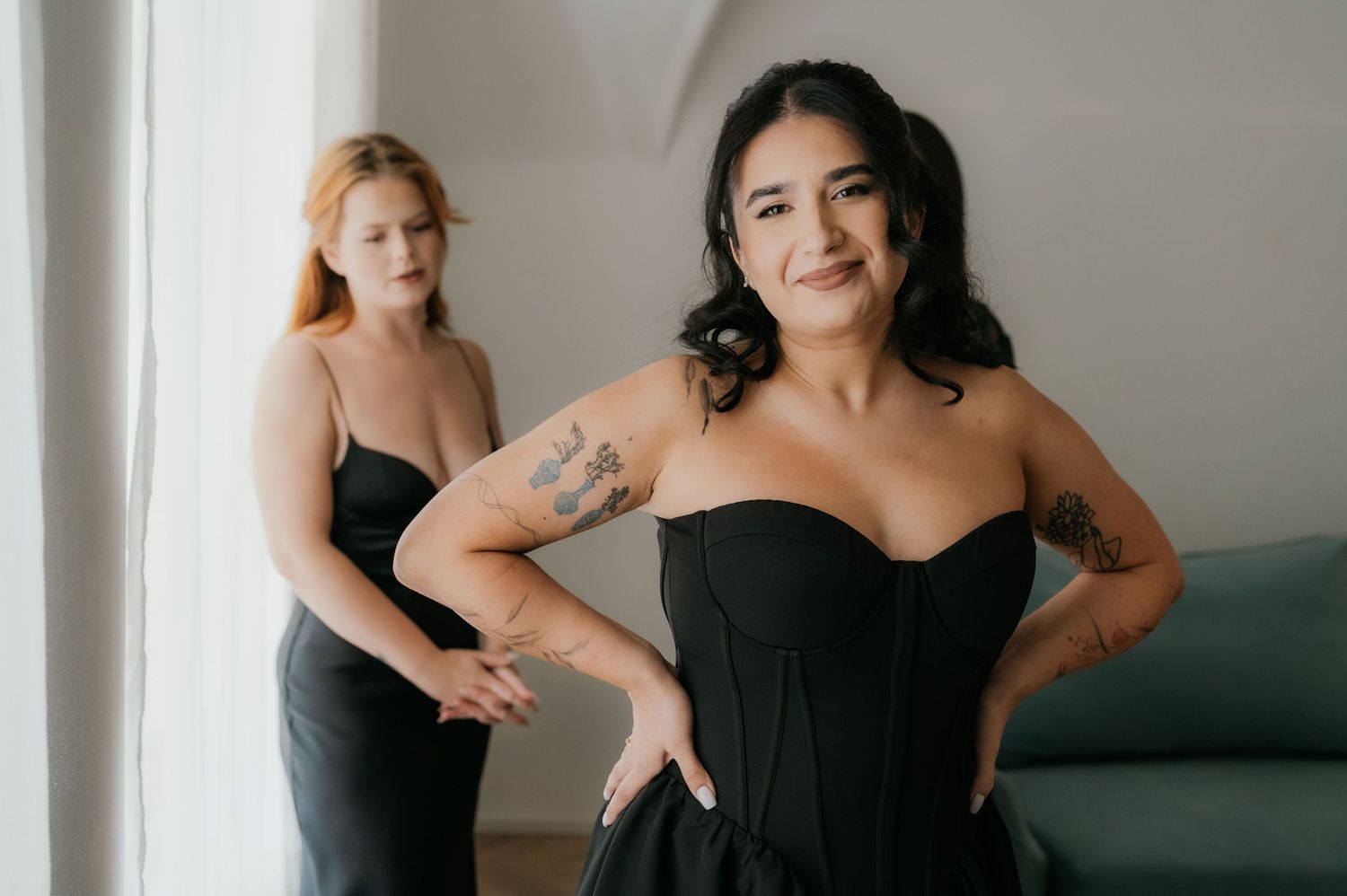 Two women in black dresses pose elegantly indoors, one with tattoos smiling confidently in a strapless gown.