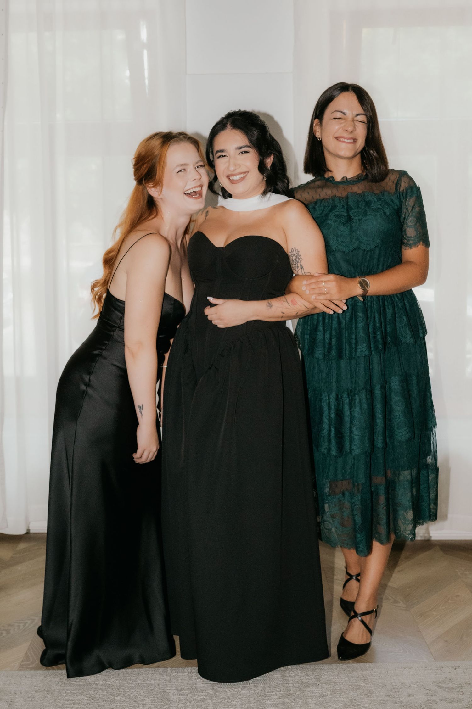 Three women in elegant black and green formal dresses smile together at an indoor event.